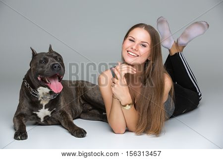 Beautiful sporty young woman lying on floor with adult grey amstafford terrier dog. Studio shot over gray background. Copy space.
