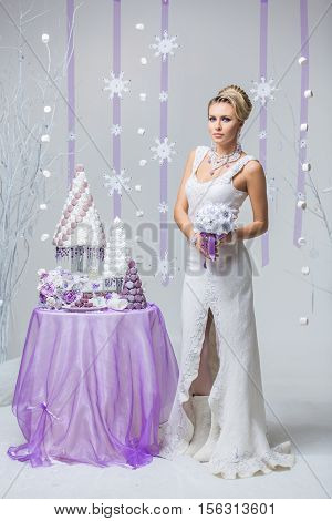 Beautiful young woman in wool wedding dress holding bridal bouquet standing near small table with big wedding croquembouche cake. Winter bride on snow flakes background. Copy space.