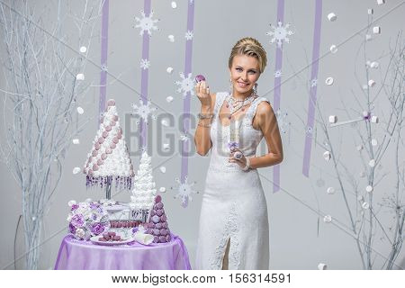 Beautiful young woman in wool wedding dress holding bridal bouquet and macaroon standing near small table with big wedding croquembouche cake. Winter bride on snow flakes background. Copy space.