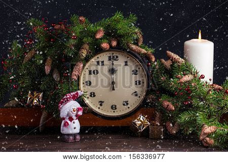 Happy New Year decoration. Vintage clock showing twelve. Candle and fir branch on wooden background. Snow flakes.