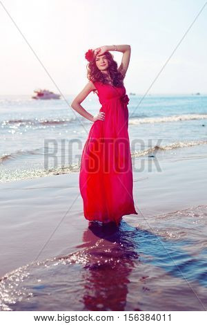 Beautiful girl in a bright red dress by the sea. Against the background of the summer lanshaft. Young romantic woman in the sunshine, sunset.
