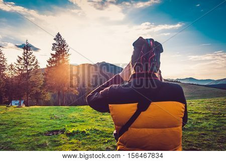 Nature photographer tourist with camera taking a photo in the mountains. Dreamy sunset landscape, spring green meadow and mountain top in the bsckground. Back view