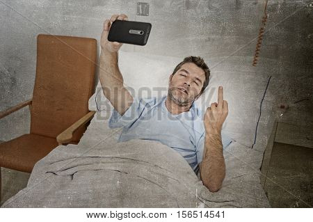 young attractive man lying on bed hospital clinic holding mobile phone taking self portrait selfie photo in pesimist recovery concept feeling angry and upset giving finger