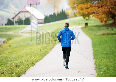 Young athlete in blue jacket running outside in colorful sunny autumn nature on an asphalt path leading through green grass. Trail runner training for cross country running.
