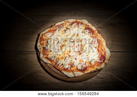Delicious round pizza with salami and pickles served on a wooden plate on old wood table in the dark