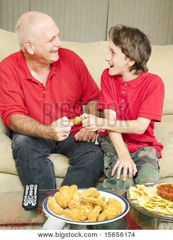Father and son playfully fighting over a chicken wing as they watch the football game.