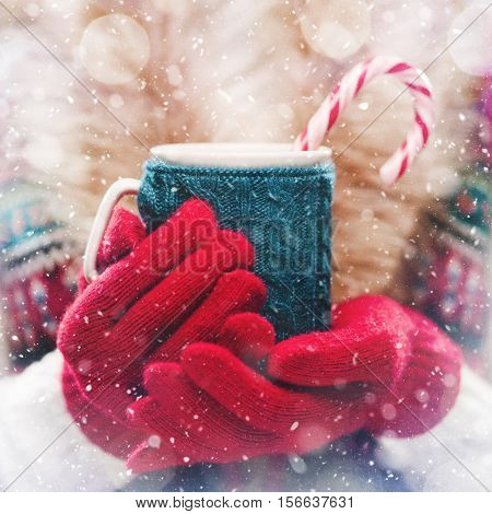 Woman holds a winter cup close up. Woman hands in woolen red gloves holding a cozy mug with hot cocoa, tea or coffee and a candy cane. Winter and Christmas time concept.