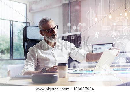Concept of digital screen with virtual icon, diagram, graph and interfaces.Adult professional stock trader wearing a classic glasses working at the wood table in modern coworking studio.Horizontal