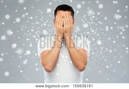 people, fear, emotions, winter and stress concept - man in white t-shirt covering his face with hands over snow on gray background