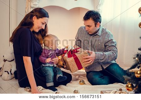 Happy family in Christmas time open gifts and presents near the Christmas tree. Happy young mother and father with their sweet baby. Merry Christmas and Happy New Year. Baby first Christmas.