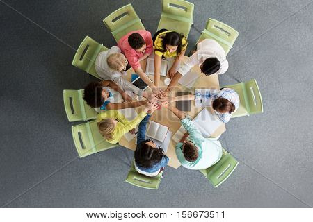 education, school, teamwork and people concept - group of international students with books and tablet pc computers putting hands on top of each other sitting at table