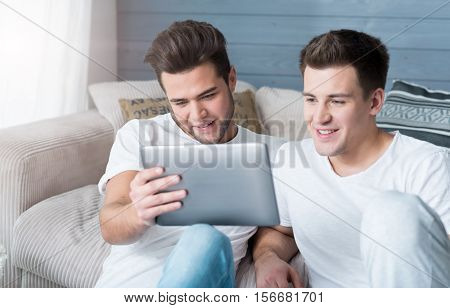 Pleasure for two. Happy cute gay couple sitting on the flor and smiling while using tablet.
