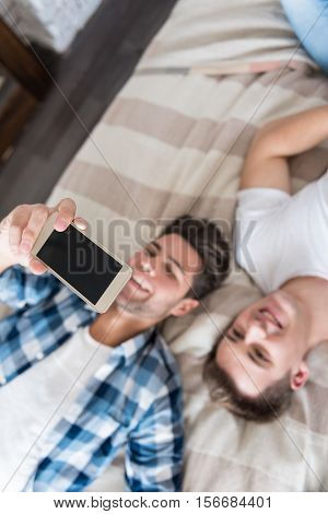 Say cheese. Smiling young gay couple making selfie and lying in bed while spending time together.