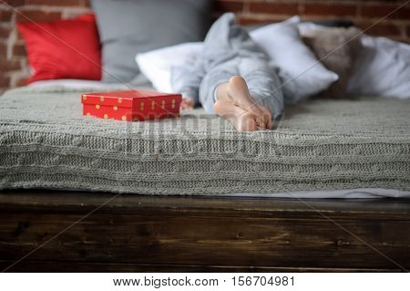 The child in pajamas lies on the big bed with his face buried in the pillow. Nearby there is a bright red box with a gift. The child isn't happy. He is disappointed with a gift and doesn't want to look at this gift. Offense. Protest.