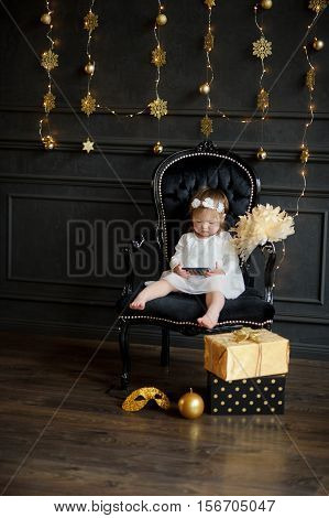 Little girl sits in chair and with enthusiasm looks at smartphone screen. With a wreath on head and barefoot baby is similar to an angel. Room is festively decorated. On a floor boxes with gifts lie.