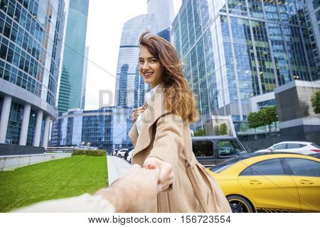 Follow Me. Young beautiful woman in beige coat drags her boyfriend, autumn streen outdoors