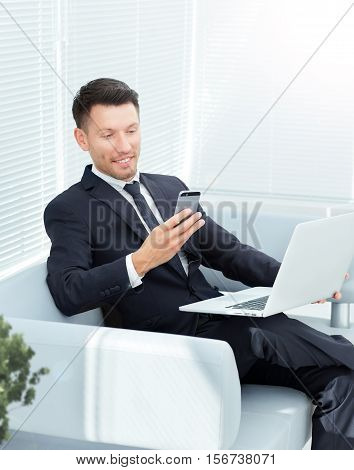 businessman sitting on sofa in the office keep a laptop open and dials the number on the smartphone