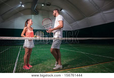 Involved in talk. Positive delighted tennis players holding rackets and talking while standing in indoor tennis court
