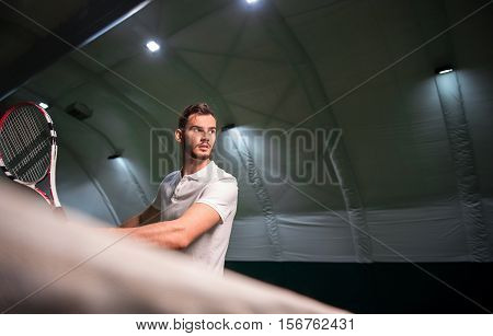 Sport ambitious. Pleasant concentrated handsome sportsman holding tennis racket and going to drive the ball while trainin gin in indoor tennis court