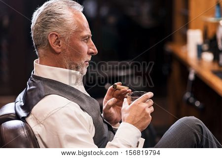 Showing status. Side view of handsome senior man wearing luxury suit sitting on armchair with cognac glass and cigar.