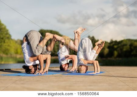 yoga, fitness, sport, and healthy lifestyle concept - group of people making tripod egg pose n mat outdoors on river or lake berth