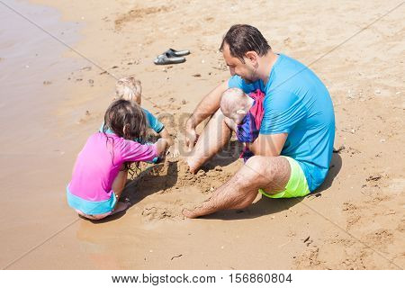 Kids burying dad in sand. UV and sun protection rashguards on the father and children. Games on the beach, sunshine