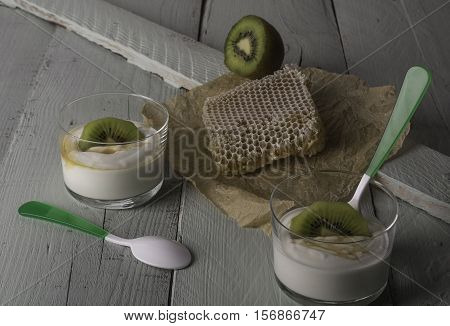 Two glasses with greek yogurt with kiwi and honey with a piece of bee panel and half a kiwi on a wooden board.
