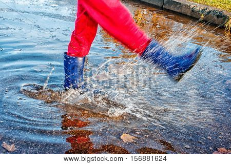 girl in blue boots and red pants standing in a puddle of water splashes with fallen yellow leaves on autuman day closeup