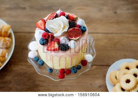 Cake with various berries, meringues and white rose on cakestand, jam cookies and tubes of pasty laid on table. Studio shot on brown wooden backbround.