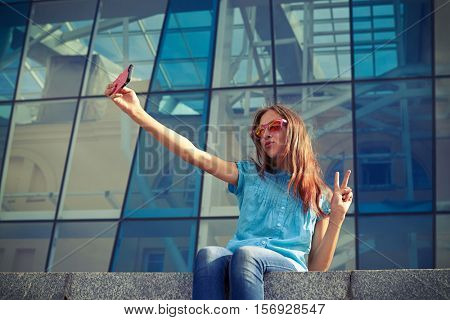 Pretty girl puckering lips while taking selfie and showing ok, sitting on the concrete bench in the downtown