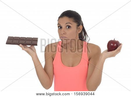 young attractive hispanic woman in fitness top holding apple fruit and chocolate bar in her hands in diet dilemma sweet tasty unhealthy food against healthy nutrition choice