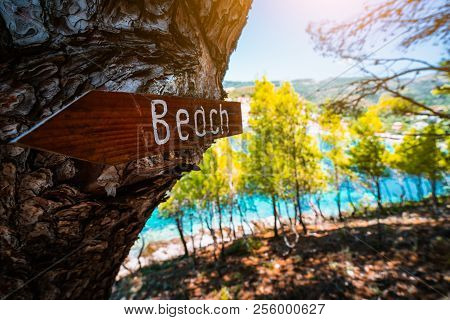 Assos Village In Morning Light, Kefalonia. Greece. Beach Wooden Arrow Sign On A Pine Tree Showing Di