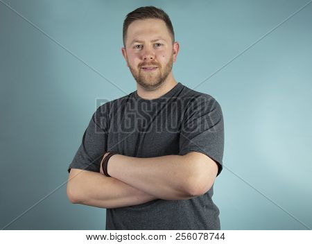 Bearded Casual Young Man In A Grey T-shirt Looking Pensively At The Camera With His With Crossed Arm