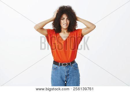 Indoor Shot Of Troubled Anxious Dark-skinned Woman With Afro Haircut Biting Lip, Holding Hands On He