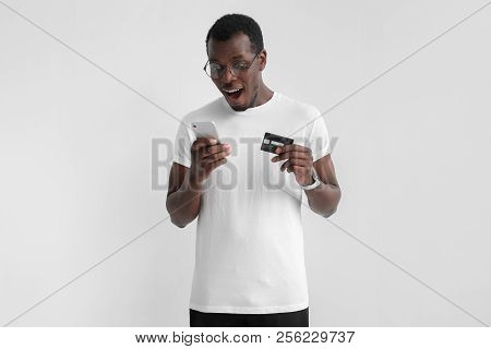 Young Handsome African American Man Standing Isolated On Gray Background, Holding Smartphone In One 