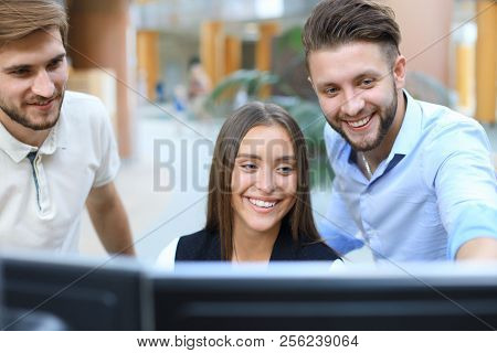 Group Of Young People In Casual Wear Sitting At The Office Desk And Discussing Something While Looki