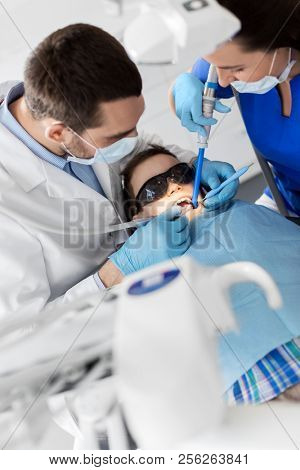 medicine, dentistry and healthcare concept - dentist and assistant with dental drill and saliva ejector treating kid patient teeth at dental clinic