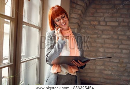 Redhead woman talking on cell phone with fascicle on workplace