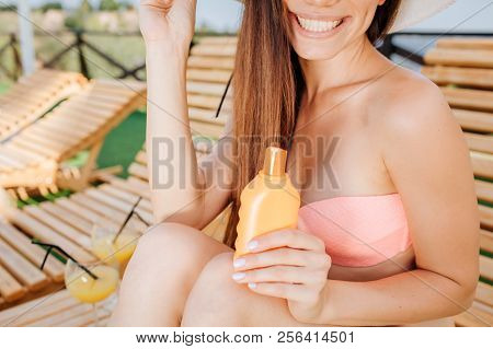 Cut View Of Girl Sits On Sunbed And Smiles. She Holds Orange Bottle In Left Hand. Girl Is Posing. Sh
