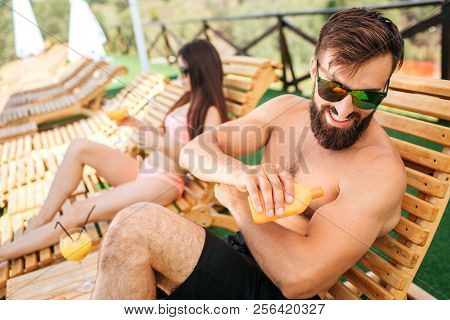 A Picture Of Bearded Guy Sitting On Sunbed And Put Some Sunproof Cream On Skin From Orange Bottle. H