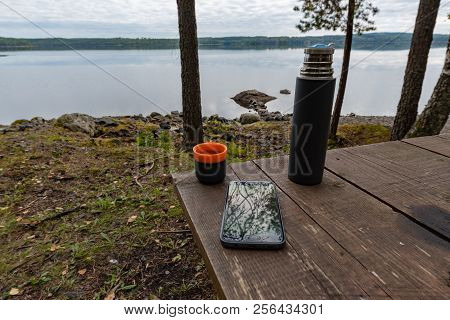 Coffeecup Thermos And A Mobile Phone At A Table Near A Lake