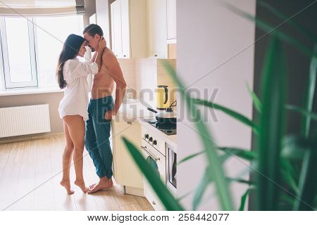 Beaytiful Man And Woman Stands In Kitchen Face To Face. She Embraces Him. Girl Stands On Toes And Ke