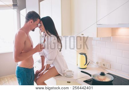 Attractive Girl Sits On Table In Kitchen And Leans To Guy. He Hodls White Cup. They Touches Each Oth