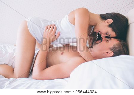 Cheerful And Nice Picture Of Couple Lying In White Bed. Girl Is Standing On Knees Above Guy And Keep