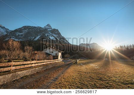 Early Winter Landscape With Dried Grass, Leafless Trees, The Austrian Alps Mountains And A Bright Su