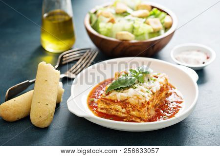 Traditional Lasagna On White Plate With Caesar Salad And Breadsticks