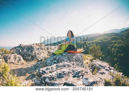 A Woman Is Sitting In A Sleeping Bag At Sunset. Camping On Top Of A Mountain. A Smiling Girl Rests A