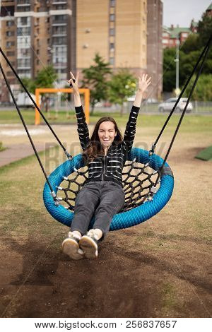 Frontal Portrait Of Young Woman Swinging On Hanging Swing Hands Up Smiling, Concept Of Freedom, Day 