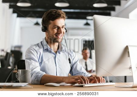 Photo of businesslike man 20s wearing office clothes and headset working on computer in call center