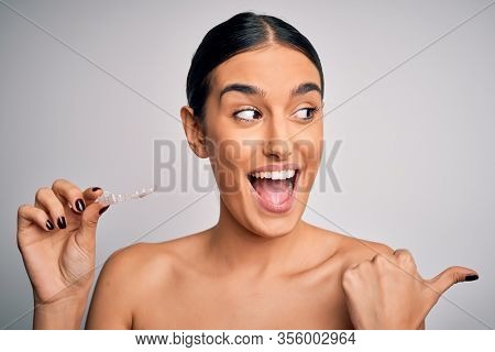 Young beautiful brunette woman holding dental aligner orthodontic to teeth corretion pointing and showing with thumb up to the side with happy face smiling